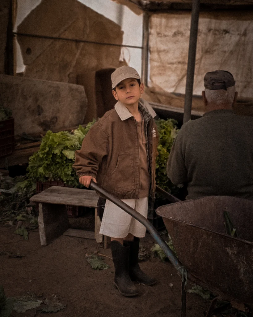 A young boy stands inside a dim greenhouse, holding the handle of a weathered wheelbarrow. He wears a brown jacket, cream shorts and rubber boots, and looks directly into the camera. Behind him, an older man sits with his back turned. Stacked green lettuce and scattered leaves fill the background.