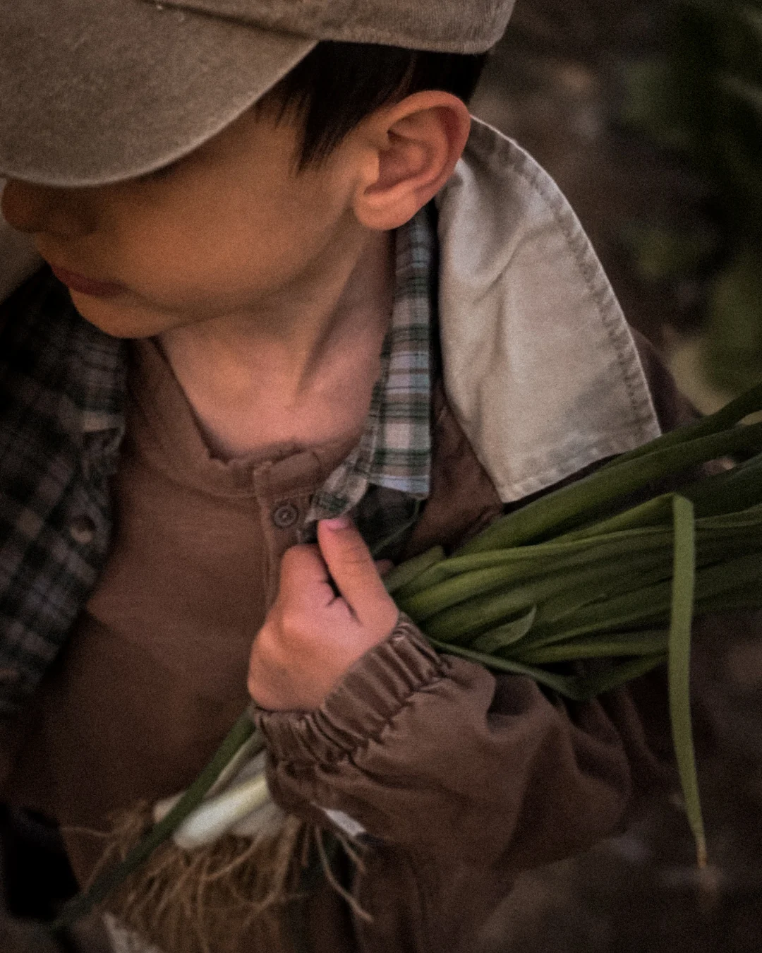 Close-up of a young boy's profile, his cap pulled low over his face. He holds a bunch of spring onions pressed tightly against his chest, one small hand gripping the stems near the white bulbs. He wears a brown jacket layered over a plaid flannel shirt. The gesture is quiet and tender — the way a child holds something they have decided matters.
