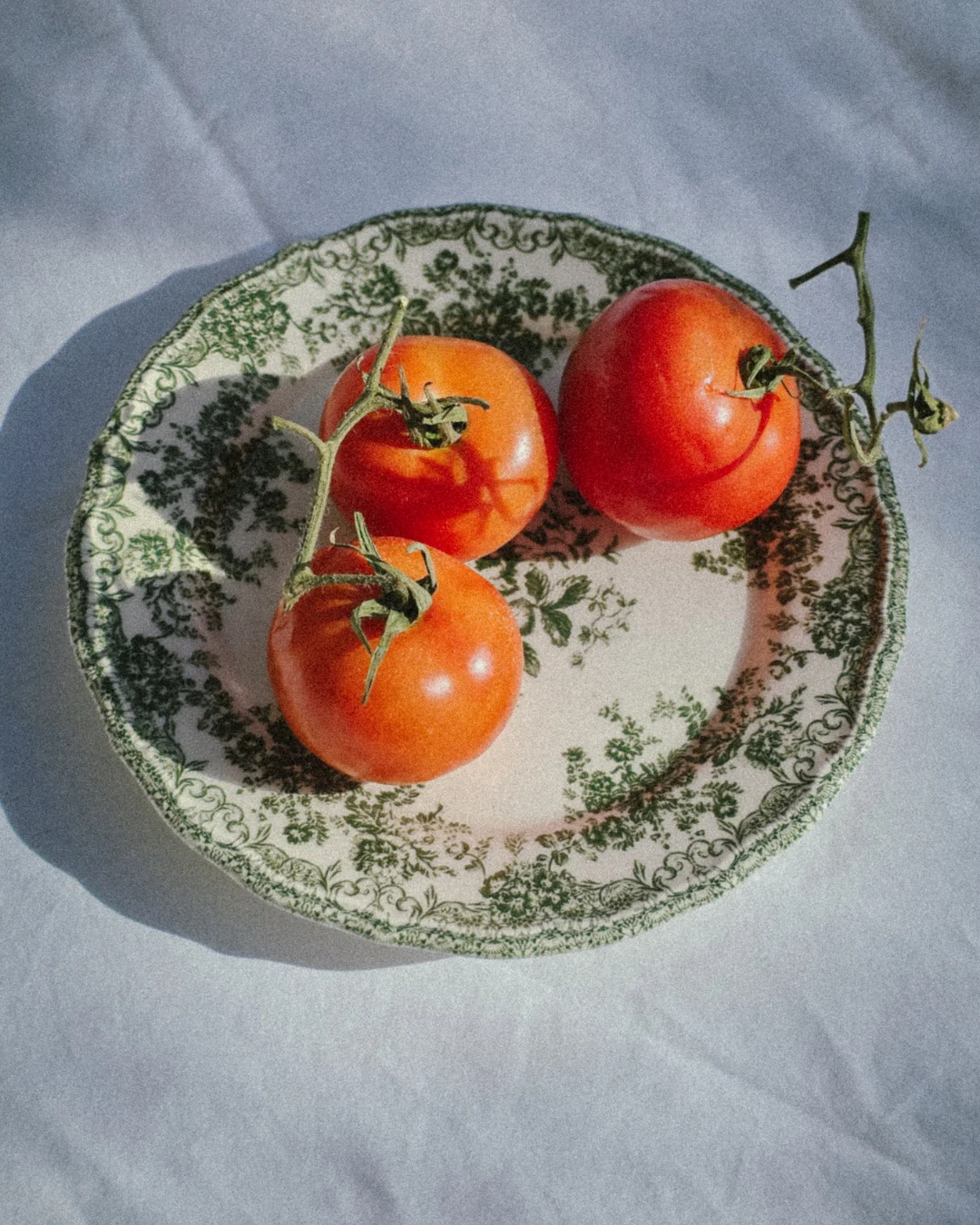 From Earth to table (16) Three ripe tomatoes with stems on a vintage green floral ceramic plate over white linen cloth in natural sunlight, farm to table aesthetic.