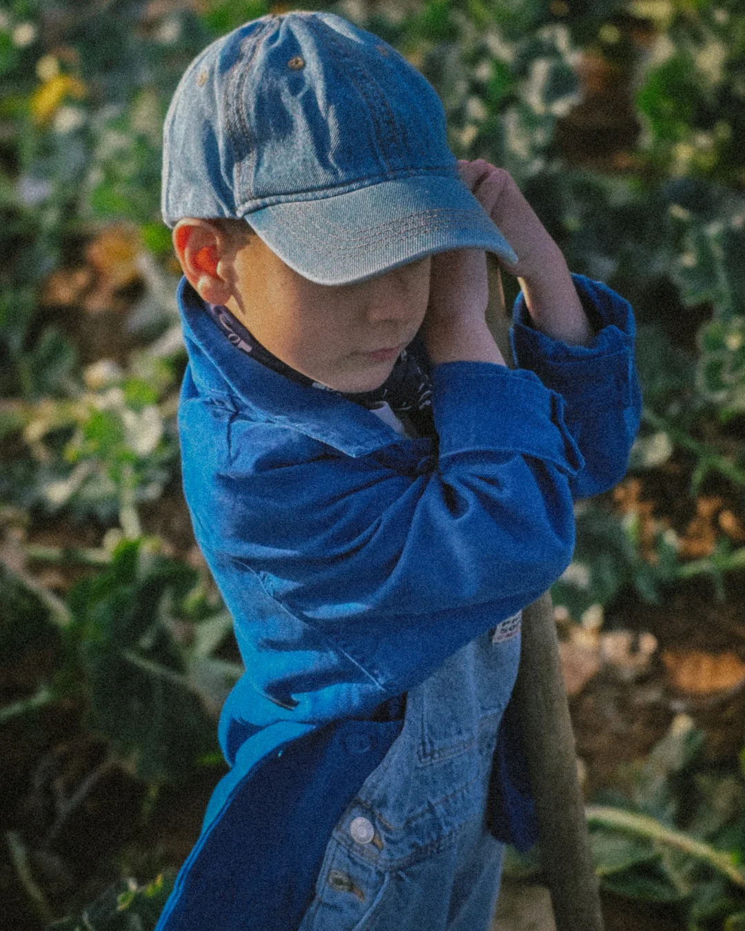 From Earth to table (27) Young child standing in a vegetable field during harvest season, wearing a blue jacket and denim cap while holding a wooden farming tool; natural sunlight, rural family farming lifestyle, slow childhood connected to the land.