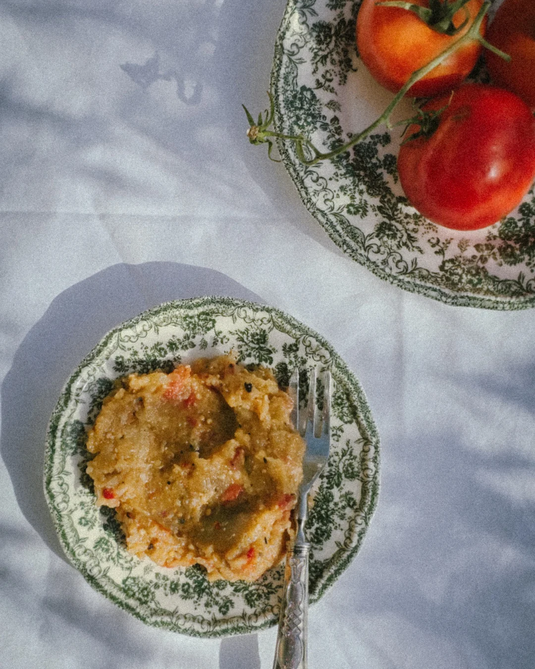 From Earth to table (6) Two vintage green floral plates on white linen outdoors, one with a traditional vegetable dish and the other with fresh tomatoes, natural sunlight casting soft shadows.