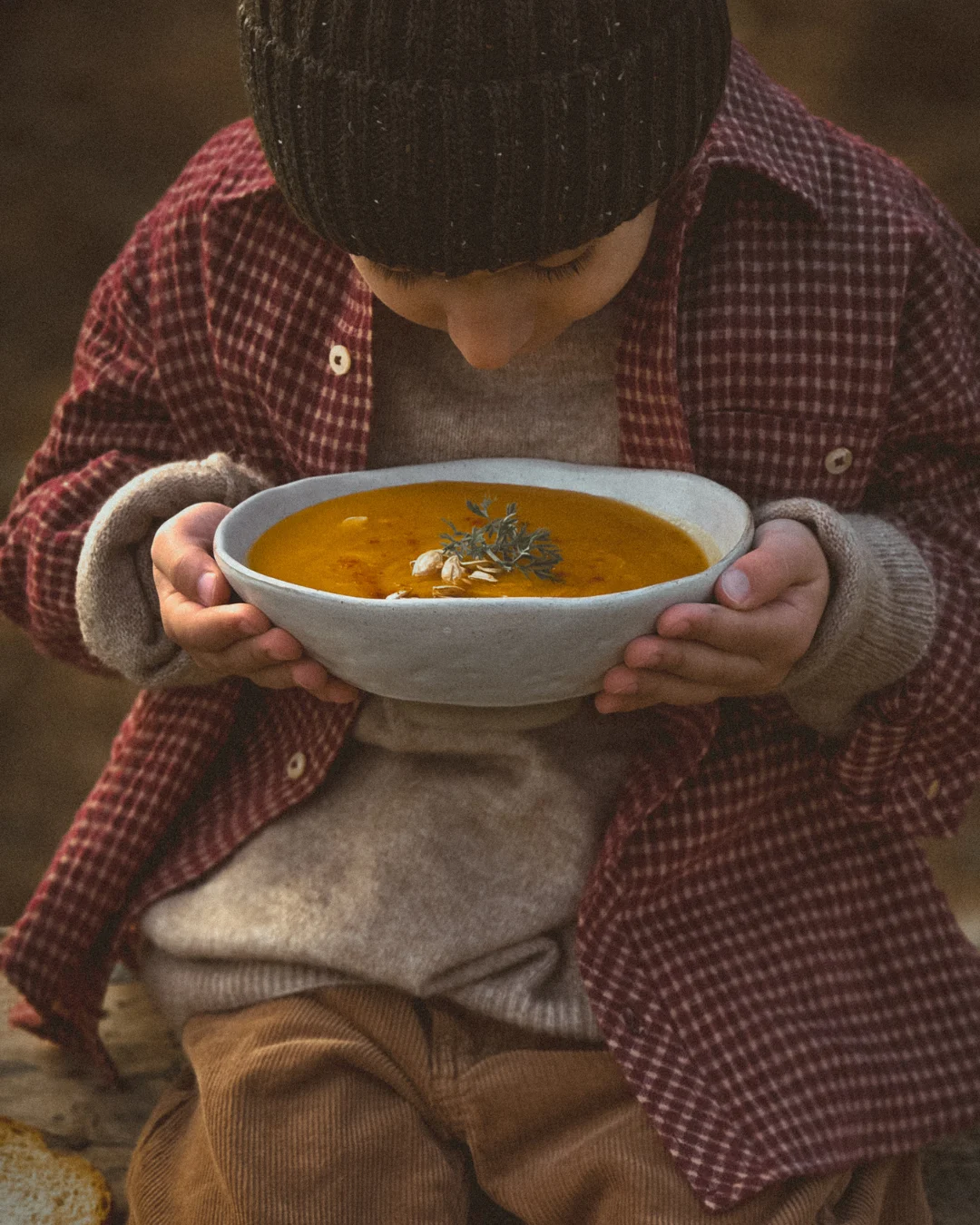 A child holding a warm bowl of homemade vegetable soup outdoors, wearing earthy clothes, captured in soft natural light, expressing a calm and cozy farm-to-table moment.