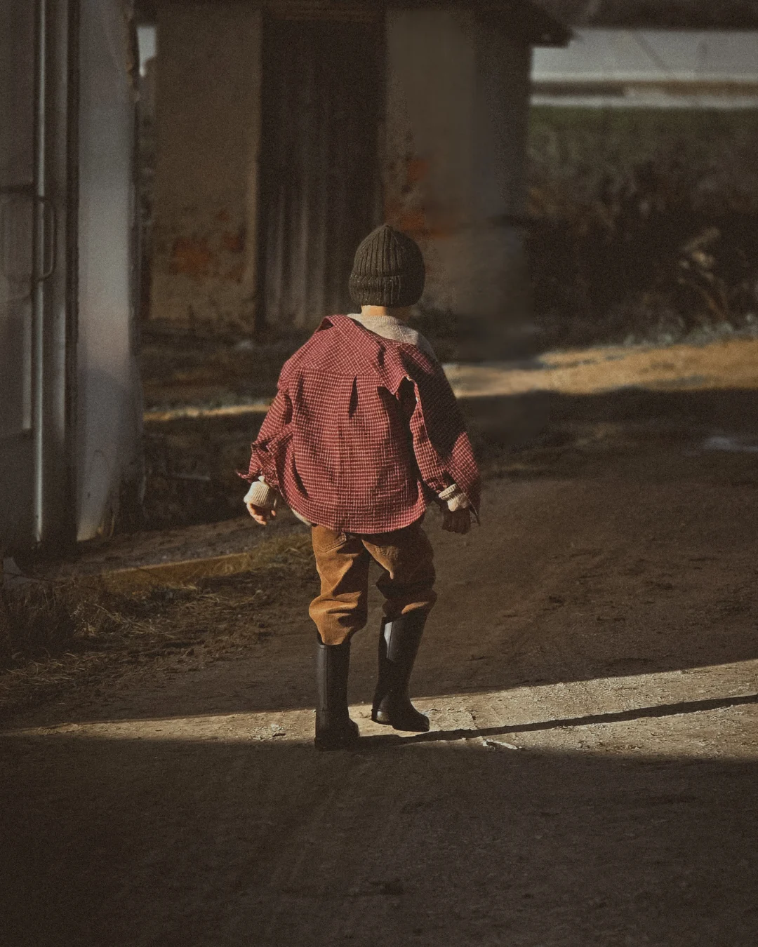 Rear view of a child walking slowly along a dirt path, wearing boots, with light and shadow stretching across the ground.