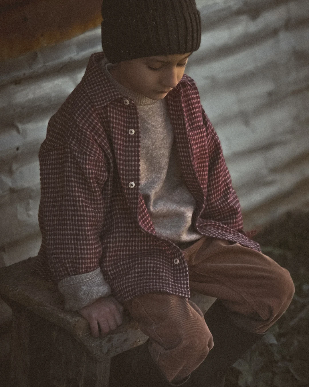 A young child sitting quietly on a low wooden surface, wearing a knit hat and layered clothes, looking down in soft evening light inside a farm setting.
