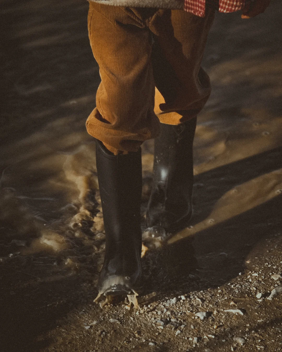 Child wearing rubber boots stepping into shallow water and soil, creating small splashes in soft evening light.