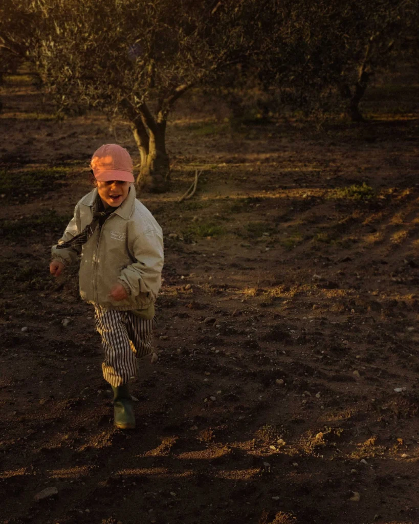 Young child running through an olive grove at golden hour, earthy ground and long shadows creating a cinematic rural atmosphere.
