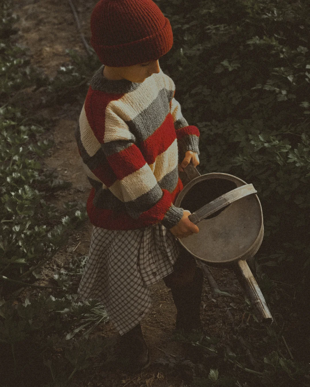 A child in a cozy striped sweater and boots holds a metal watering can while walking through a patch of green plants.