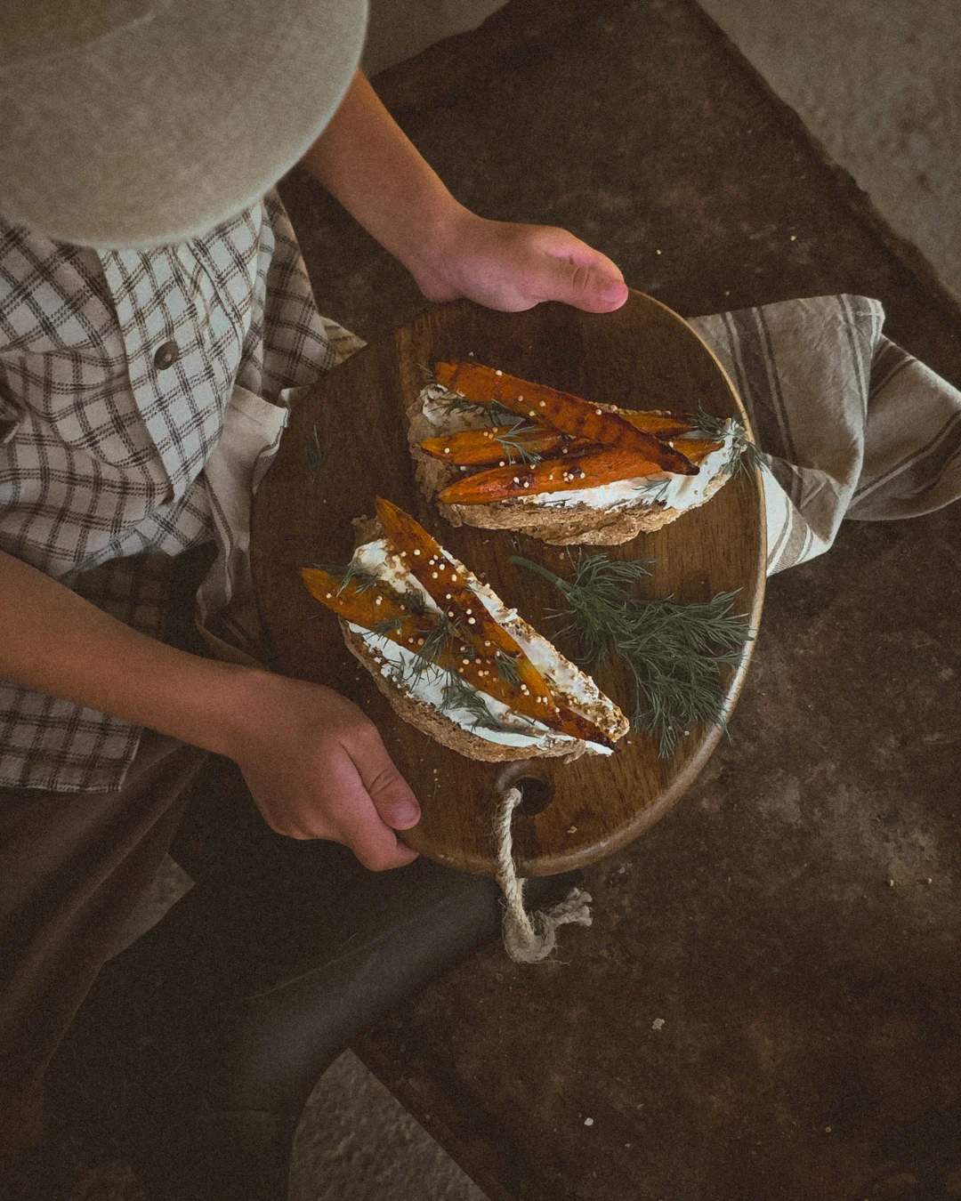 A child holding a wooden board with rustic bread topped with roasted carrots, cream cheese, and fresh dill in warm, earthy light.