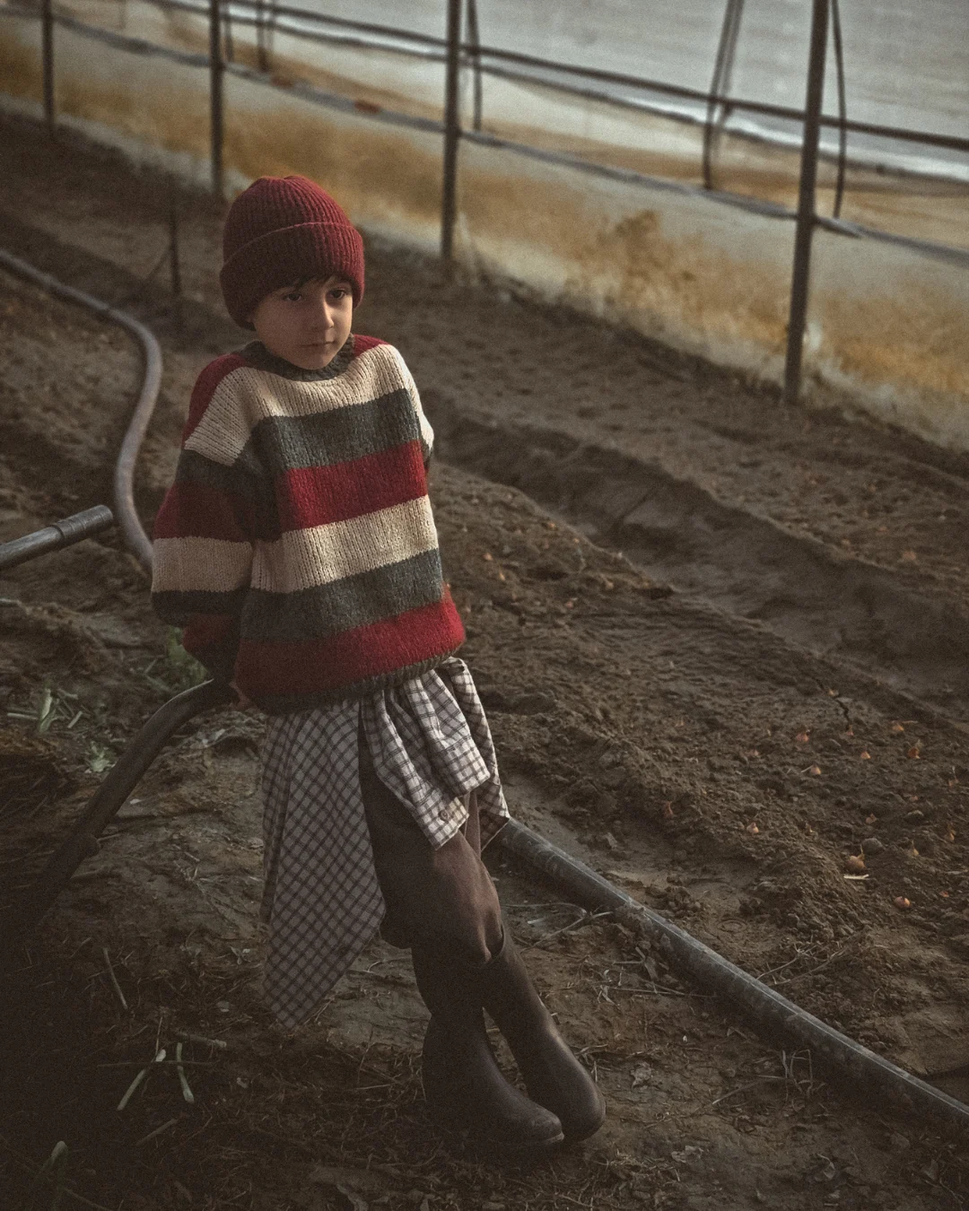 A young boy wearing a striped winter sweater and red beanie stands inside a dimly lit greenhouse, leaning on irrigation pipes. Earthy tones, cinematic shadows, early winter farm atmosphere.