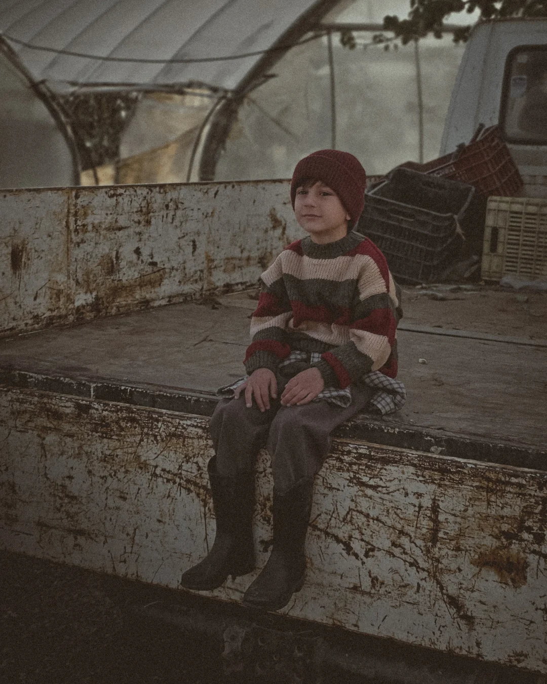 Young child wearing a striped knit sweater and beanie sitting on the back of an old pickup truck, with crates and greenhouse structures in the background.