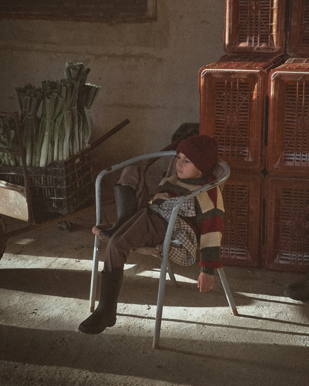 A young child wearing a striped sweater, brown pants, and a red beanie sits thoughtfully on a metal chair inside a rustic farm shed with crates and freshly harvested leeks behind them. Soft winter light falls across the scene