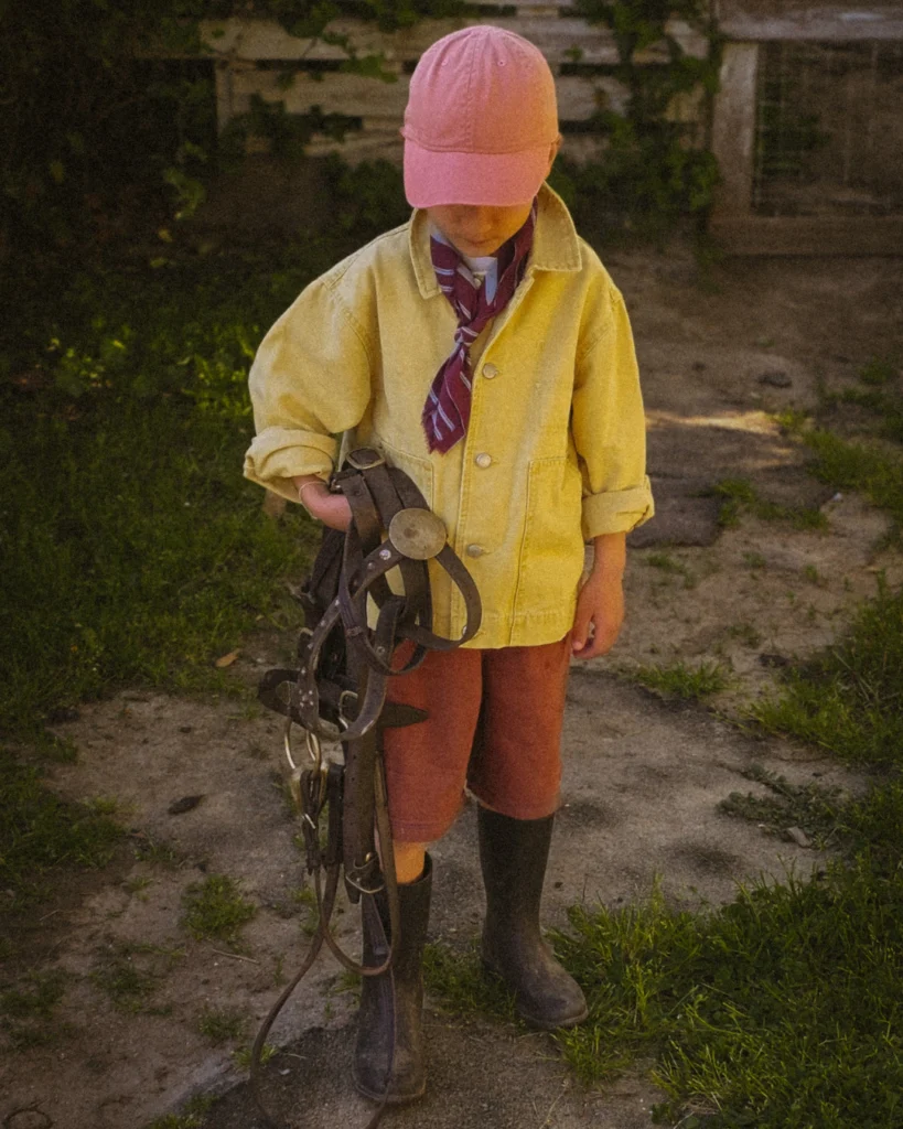 A young boy stands alone on a dirt path, head bowed, holding a heavy leather bridle with both hands. He wears a yellow linen jacket, rust-colored shorts, tall dark boots and a pink cap with a burgundy scarf at his neck. The light is dim and dramatic, falling from above. Dense green ivy and dark wooden boards frame the background. The mood is still and weighted.