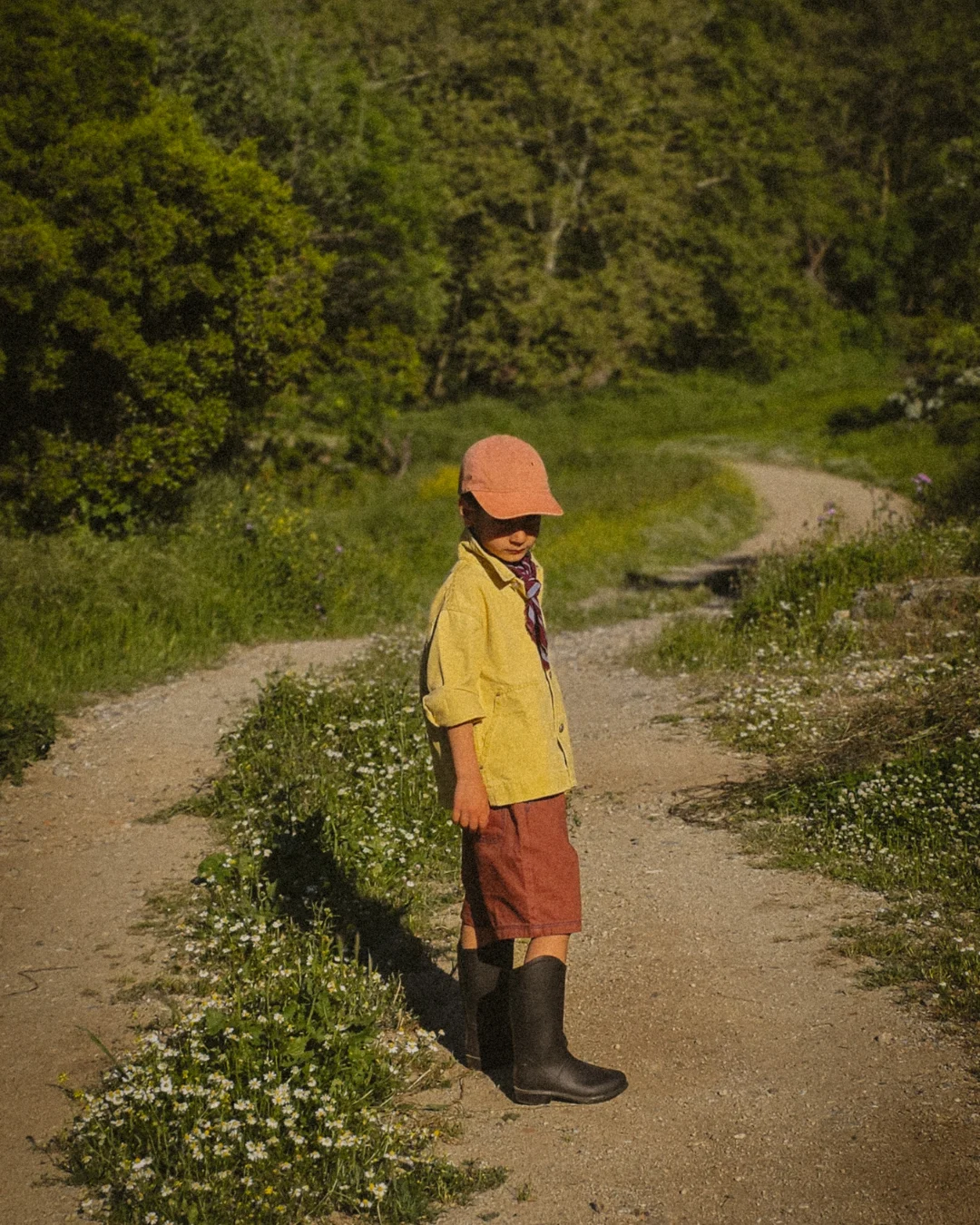 A young boy stands alone on a narrow dirt path, head bowed, eyes downward. He wears a yellow linen jacket, rust-colored shorts, tall dark boots and a dusty pink cap with a burgundy scarf at his neck. Small white wildflowers line the edges of the path. The road curves and continues behind him into a dense wall of green trees. The light is warm and dappled. The mood is still, reflective, and quietly conclusive.